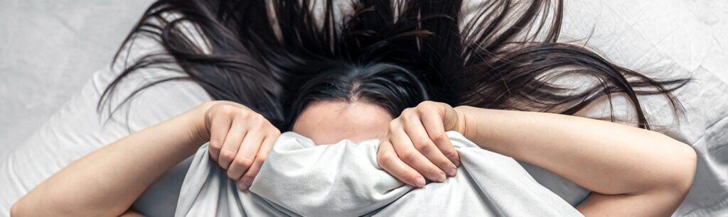 Bright young woman covering her face with her arms lying on a bed, illustrating the importance of mental health support and the role of The Study Buddy website in providing effective help.