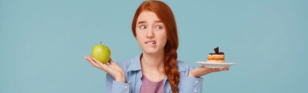 Unsure whether to choose healthy or indulgent snacks, young woman with red hair holds an apple and cake, illustrating the importance of balanced choices with The Study Buddy website.