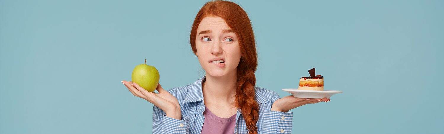Unsure whether to choose healthy or indulgent snacks, young woman with red hair holds an apple and cake, illustrating the importance of balanced choices with The Study Buddy website.